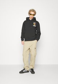 Felpa nera con cappuccio e stampa grafica, pantaloni cargo beige con più tasche e sneakers nere con strisce bianche. Occhiali da sole gialli.