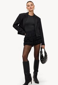 Vrouw in een zwarte cropped blazer, met gerimpelde shorts, doorschijnende panty, kniehoge laarzen, ringoorbellen, en houdt een gestikte zwarte handtas vast.