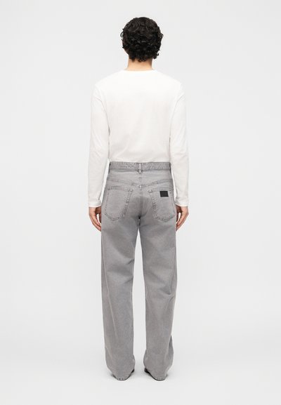 Άνδρας με σγουρά μαλλιά στέκεται με την πλάτη του γυρισμένη, φοράει λευκό πουκάμισο με μακριά μανίκια και φαρδιά γκρι τζιν, σε απλό φόντο.