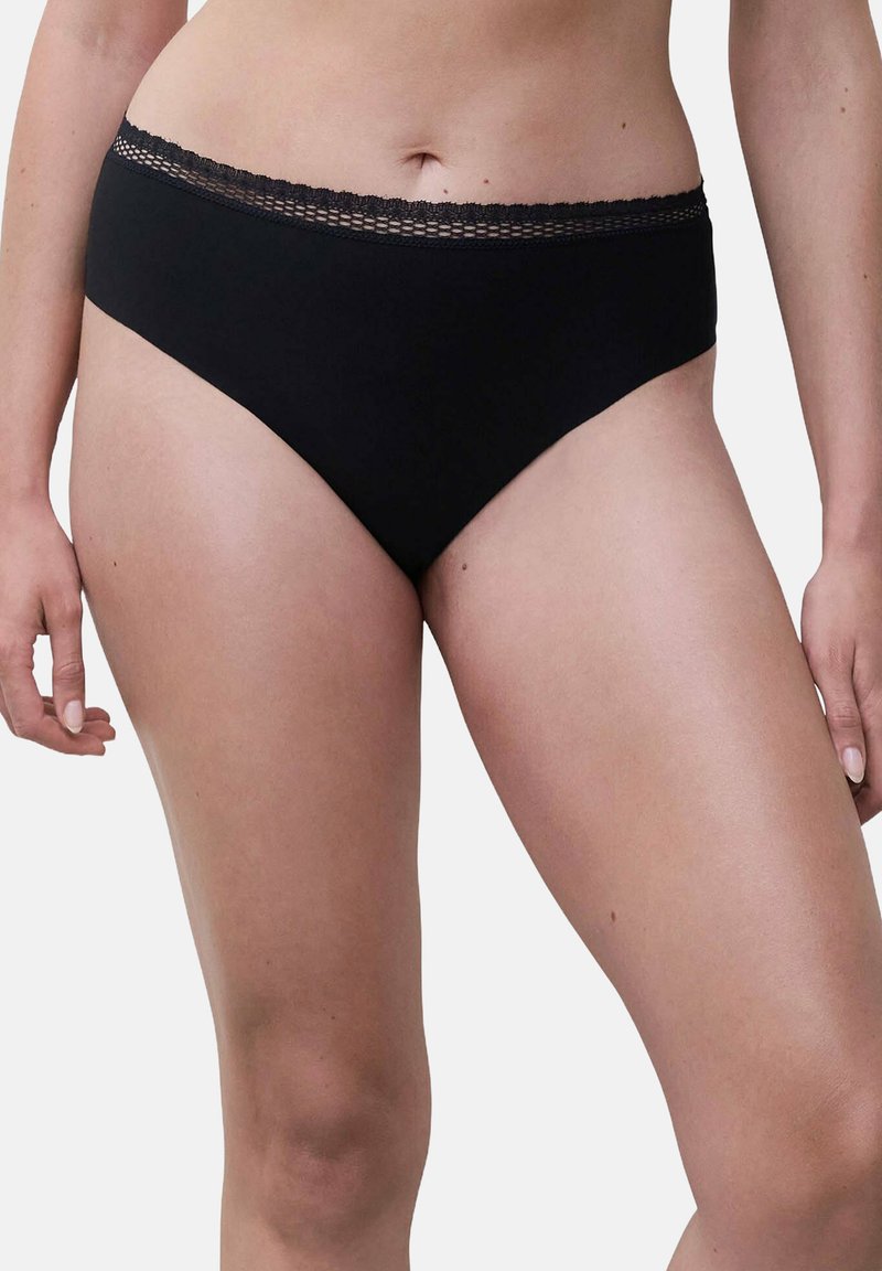 Frau, die schwarze Unterwäsche mit mittlerem Bund und Spitzendetails trägt, von der Taille bis zu den oberen Oberschenkeln vor einem hellen Hintergrund gezeigt.