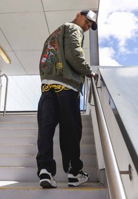 Chaqueta bomber verde con bordado colorido de dragón, jeans negros de corte suelto con acentos de logotipo, y zapatillas en blanco y negro sobre los escalones.