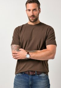 Muž s vousy, oblečený v hnědém tričku a džínách, se zkříženýma rukama, které odhalují tetování a náramkové hodinky s hnědým koženým řemínkem.