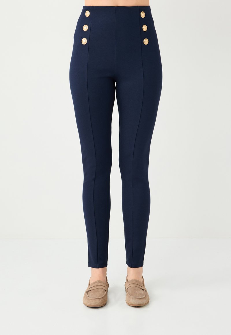 Pantalon taille haute bleu marine en tissu extensible, avec six boutons décoratifs dorés le long de la taille et des coutures verticales pour une silhouette ajustée.