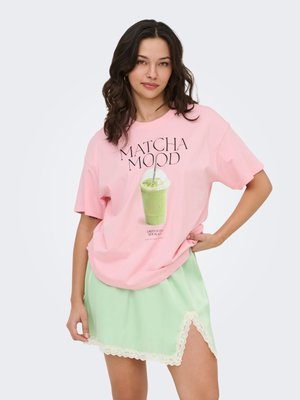 Kvinde iført en lyserød oversized T-shirt med teksten "MATCHA MOOD" og en grafik af en grøn drik, kombineret med et lysegrønt skørt med blondekant.
