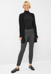Zwarte lange cardigan over een zwarte coltrui, gecombineerd met grijs gestreepte slim-fit broek en zwarte instappers. Soepele texturen en een minimalistisch ontwerp.