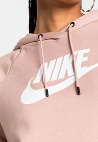 Rosa hoodie i mjukt material, med en vit Nike-logotyp. Den har en framficka i kängurustil och en huva med dragsko med svarta toppar.