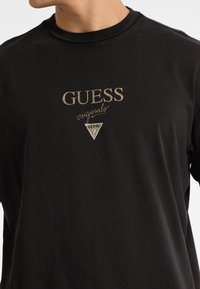 T-shirt a maniche lunghe nera con logo "GUESS originals" in oro e argento e simbolo triangolare sul davanti. Materiale in cotone morbido.