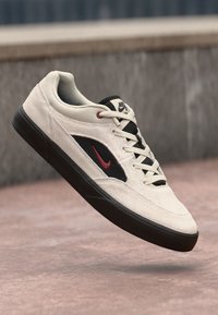 Μπεζ σουέτ αθλητικά παπούτσια με μαύρη πλαϊνή πάνελ, κόκκινο σήμα τικ και μαύρη καουτσούκ σόλα. Διαθέτει σχέδιο με κορδόνια και επενδεδυμένο κολάρο.