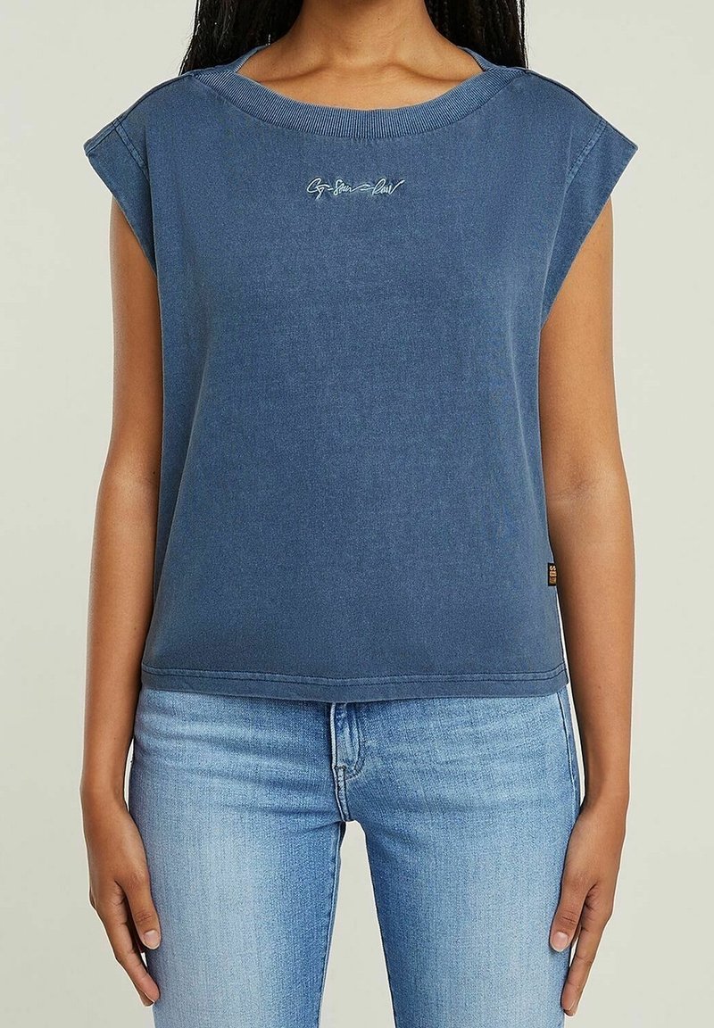 Žena v bezrukávovém modrém topu s kulatým výstřihem a malým bílým textem na prsou, v kombinaci se světle modrými džíny.
