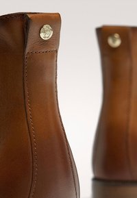 Brązowe skórzane botki za kostkę z gładką fakturą, zaokrąglonym kształtem oraz metalicznym guzikiem przy górze.