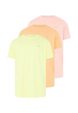 Mężczyzna z brodą o muskularnej budowie, ubrany w jasnżółtą koszulkę z krótkim rękawem i niebieskie dżinsy, stojący na jednolitym jasnym tle.