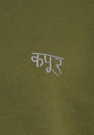 Tessuto verde oliva con una texture liscia, caratterizzato da testo ricamato bianco in una calligrafia stilizzata sul lato sinistro superiore.
