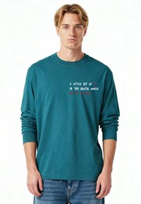 T-shirt à manches longues couleur sarcelle en coton, avec texte blanc et rouge affichant un message sur le devant. Col rond simple et coupe décontractée.