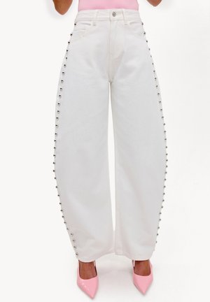 Pantalones blancos de pierna ancha con tachuelas plateadas a lo largo de las costuras exteriores, combinados con tacones rosa de punta puntiaguda y un top rosa claro.