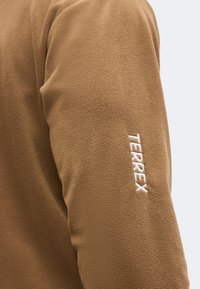 Manche en polaire marron avec logo "TERREX" blanc brodé sur le haut du bras, sur un fond clair uni.