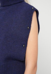 Suéter em azul-marinho, tricotado, com gola alta, sem mangas e botões pretos decorativos no ombro. Tecido texturizado com bainha canelada.