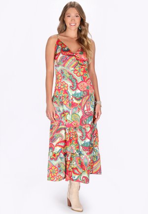 Egy nő áll, mosolyog, színes virágos paisley mintás maxi ruhát visel vékony pántokkal és bézs bokacsizmával.