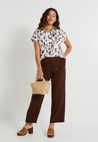 Chemise à manches courtes florale en blanc, marron et noir ; pantalon large marron ; sandales à plateforme beiges ; tenant un petit sac en osier.