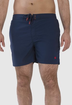 Mann trägt dunkelblaue Badeshorts mit Kordelzug in der Taille und steht vor einem schlichten Hintergrund.