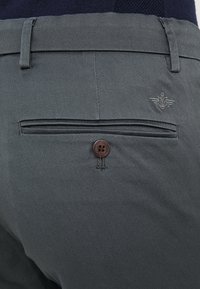 Mörkgrå byxor med en bakficka med en brun knapp och subtil logotypbroderi. Materialet har en slät yta.