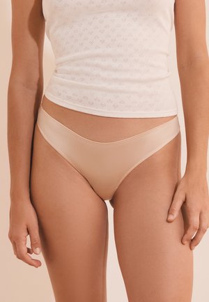 Femme portant un sous-vêtement beige taille haute et un haut blanc sans manches avec de petits motifs perforés, montrée de la mi-cuisse jusqu’à la taille.