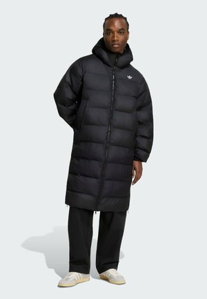 Ung person med flettet hår iført en lang sort polstret frakke, sorte bukser og lyse sneakers, der står mod en bleg baggrund.