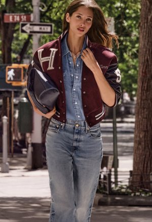 Frau geht draußen und trägt eine kastanienbraune College-Jacke über einem Jeanshemd und Jeans, hält eine dunkelblaue Tasche unter dem Arm.