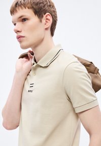 Μπεζ πόλο μπλούζα με μαύρες λεπτομέρειες, διαθέτει τρία κουμπιά μπροστά και λεπτομέρεια λογότυπου. Το μοντέλο κρατά καφέ τσάντα στον ώμο.