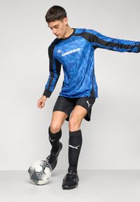 Sinine ja must pikkade varrukatega jalgpallisärk, millel on "UMBRO" logo, koos mustade lühikeste pükste ja sokkidega, kõrvala must-valge jalgpall.