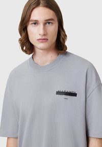 T-shirt en coton gris clair avec un col rond, des manches courtes et une étiquette graphique imprimée portant "ALLSAINTS" et du texte japonais.