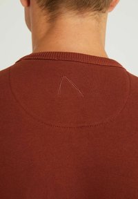 Maglione rigato di colore ruggine con scollatura rotonda, caratterizzato da un piccolo dettaglio ricamato a forma di triangolo sul retro. Il materiale sembra morbido.