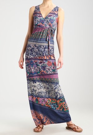 Nainen, joka pitää päällään hihattoman, monivärisen kuvioidun maksi-mekon, jossa on vyötärösolmu, ja ruskeat matalat sandaalit vaalealla taustalla.