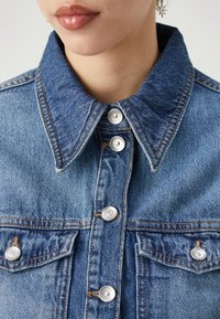 Modrá džínová bunda s výrazným límcem, stříbrnými knoflíky a dvojitými náprsními kapsami, s viditelným prošíváním a texturovaným povrchem.