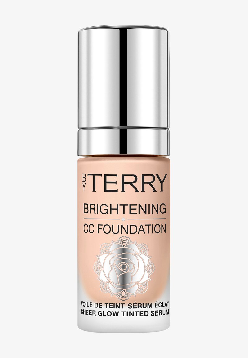 Bouteille pêche clair avec un capuchon pompe en argent, étiquetée "BY TERRY BRIGHTENING CC FOUNDATION." Présente un design de rose blanche décoratif.