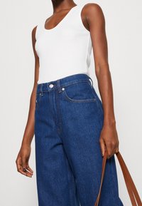 Person wearing a white sleeveless top and high-waisted blue jeans, holding brown bag straps with one hand against a plain background.