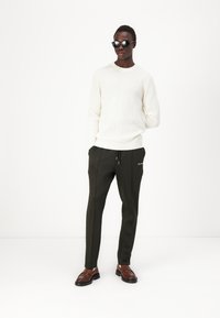 Maglione bianco a costine, pantaloni tapered verde scuro con coulisse, scarpe di pelle marrone e occhiali da sole neri. Sfondo neutro.