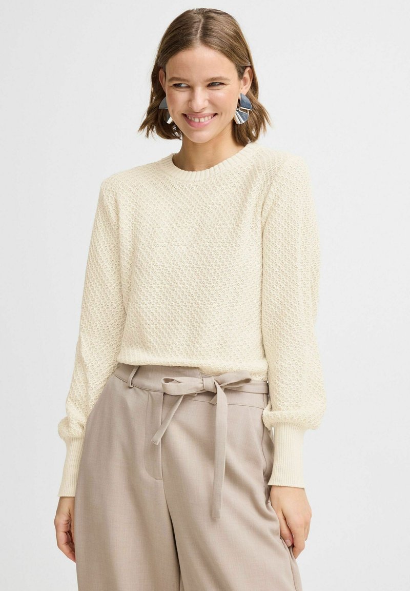 Pull crème avec un tricot texturé en motif losange, col rond et manches longues. Assorti avec un pantalon beige doté d'une ceinture nouée.