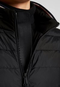 Czarna puchowa kurtka z wysokim kołnierzem, czarnym, teksturowanym zamkiem błyskawicznym oraz warstwowym wzorem materiału. Na zakończeniu zamka znajduje się subtelne logo.