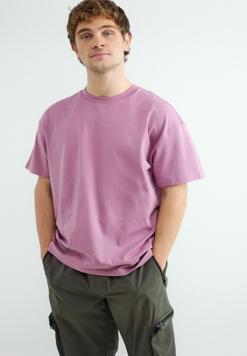 Jonge man met krullend haar, die een losse roze T-shirt en olijfgroene cargobroek draagt, handen in de zakken, tegen een effen witte achtergrond.