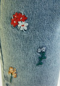 Tissu en denim avec de petites fleurs brodées en rouge, blanc, orange et violet dispersées sur la surface.