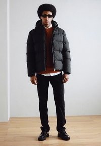 Czarna kurtka puchowa, nałożona na pomarańczowy sweter, w połączeniu z czarnymi spodniami i czarnymi butami. Model nosi ciemne okulary przeciwsłoneczne i ma fryzurę w afro.