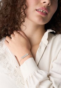 Bracciale argentato regolabile con un charm rettangolare dalla superficie liscia, indossato su un polso, con una blusa bianca a trama nel fondo.