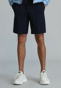 Shorts bleu marine en tissu avec des plis sur le devant, associés à des baskets blanches épaisses, avec une semelle texturée et un design à lacets.