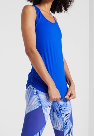 Femme portant un haut bleu sans manches et des leggings à motifs bleu et blanc sur un fond clair uni.