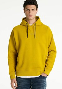 Sennepsgul hoodie med snøre i hætten, ribbede manchetter og et diskret logo på brystet. Materialet ser blødt og struktureret ud.