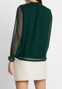 Blusa verde trasparente con maniche lunghe, scollatura arricciata e polsini arruffati; indossata sopra una gonna chiara e texturizzata.