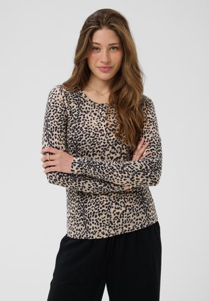 Femme aux longs cheveux bruns portant un haut à manches longues beige et noir à imprimé léopard et un pantalon noir, bras croisés, fond neutre.