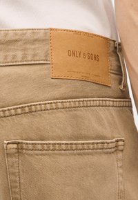 Κοντινό πλάνο τσέπης και ζώνης παντελονιού από μπεζ τζιν, με καφέ δερμάτινο ταμπελάκι "ONLY & SONS" και λεπτομέρειες ραφών.
