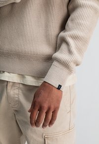 Beige gerippter Strickpullover mit strukturierten Details und einem schwarzen Stoffetikett am Bund, kombiniert mit hellen Cargo-Hosen.