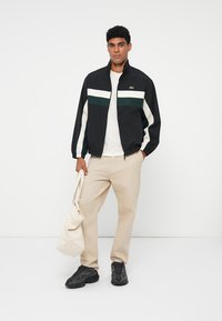 Jachetă neagră cu dungi albe și verzi, asortată cu un tricou bej și pantaloni bej deschis. Încălțăminte sport neagră și o geantă de umăr crem.